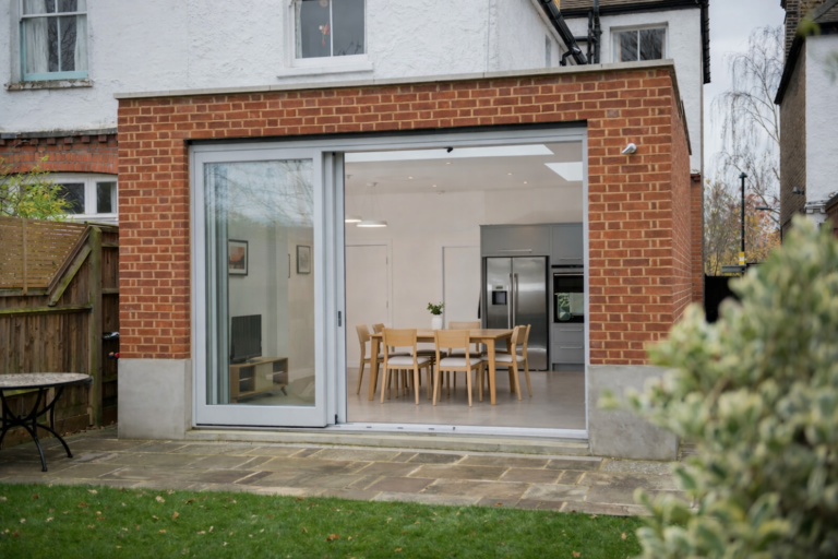 Modern York‑style home extension featuring an in‑line sliding patio door with a light‑wood Scandinavian dining area visible inside; red brick exterior, stone patio, and garden lawn in the foreground.
