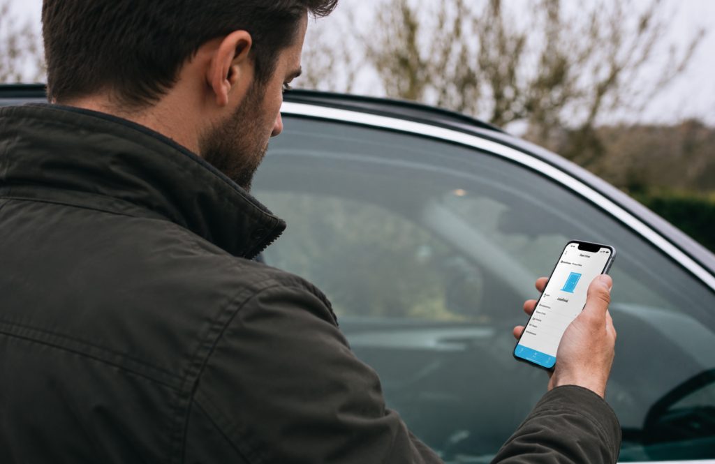 Man standing beside a car looking at a smartphone in an outdoor setting.